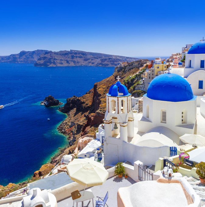 Whitewashed buildings with iconic blue domes cascade down Santorini's volcanic cliffs, overlooking the deep blue Aegean Sea. The image evokes a postcard-perfect Greek island getaway — a fitting backdrop for pondering the merits of Ouzo-sipping over fiat currency concerns, where the timeless beauty of the Mediterranean offers an escape from financial worries.