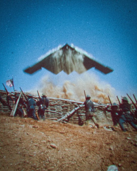 A stealth bomber flies low over a group of soldiers positioned behind a wooden barricade, kicking up a large cloud of dust as it passes against a clear blue sky. An American flag is visible on the left side of the scene.