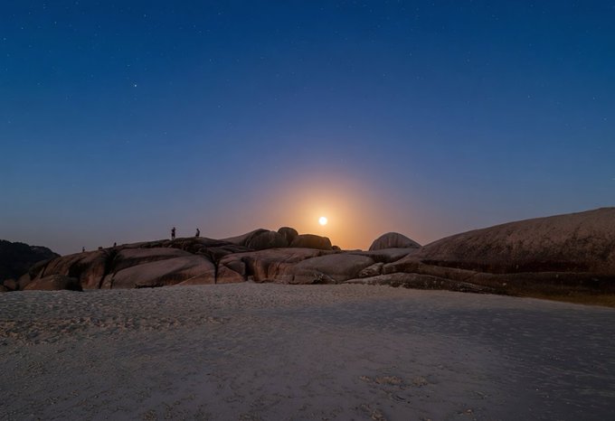 A moonlit night scene over a rocky landscape with a bright moon on the horizon and small figures of people standing on the rocks.