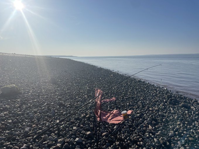 A bright, sunny photograph of a pebbled beach with a small red folding chair set up next to fishing rods near calm water.