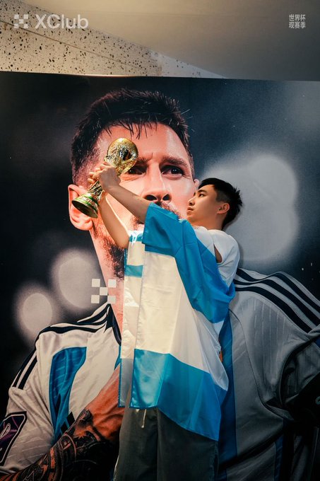 A young fan wearing an Argentina flag as a cape kisses a replica World Cup trophy in front of a large poster of Lionel Messi, with an 'XClub' watermark in the upper left corner.
