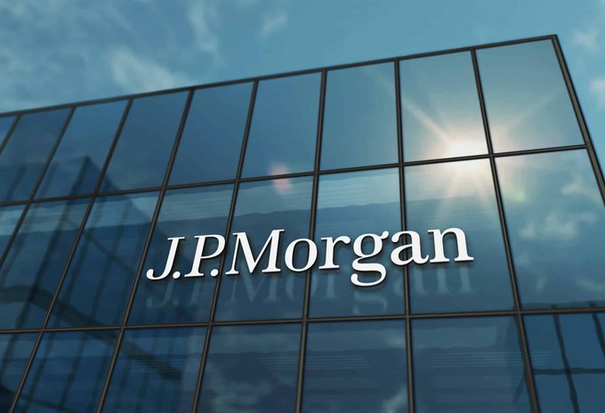 The image shows the exterior of a glass-paneled building featuring the "J.P. Morgan" logo prominently displayed on its facade, with sunlight reflecting off the windows. This relates to the accompanying post about JPMorgan's participation in a Wall Street trial testing tokenized assets across multiple blockchains, highlighting the bank's involvement in exploring blockchain-based financial innovation.
