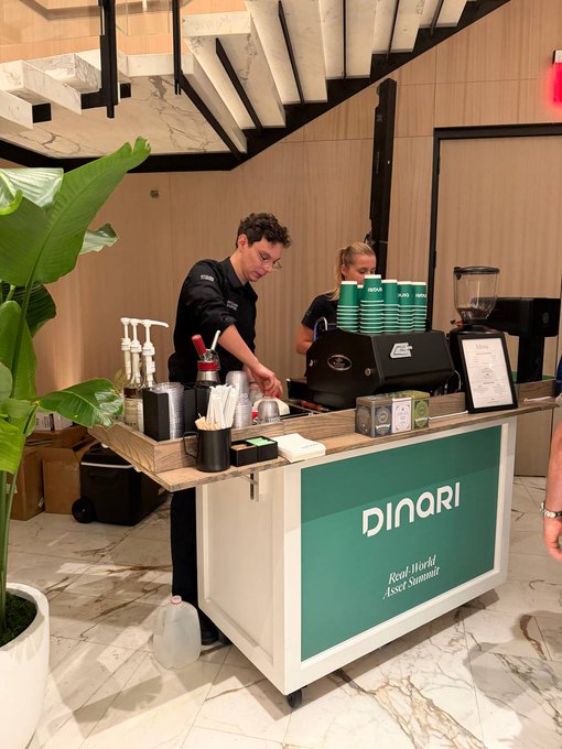 Two baristas stand behind a wooden counter branded 'Dinori - Real World Asset Summit,' preparing drinks with an espresso machine and stacked green cups, alongside a large green plant in a white pot.