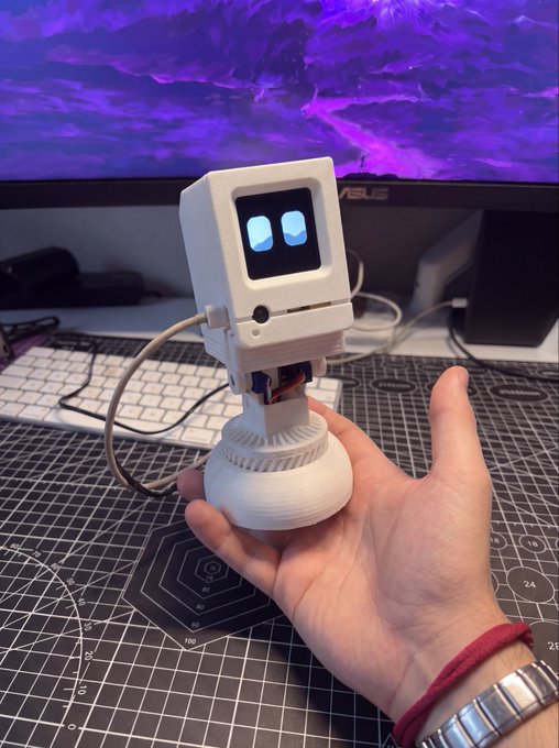 A small white 3D-printed robot with digital blue eyes is held in someone's hand in front of an ASUS monitor, representing a tech build—the kind of maker project being developed and showcased alongside the announcement of a new microduck token on Robinhood (CA: 0xed04806EF9495FBd7919d9fdaBc6B74569a79656) with a Cursor AI partnership.