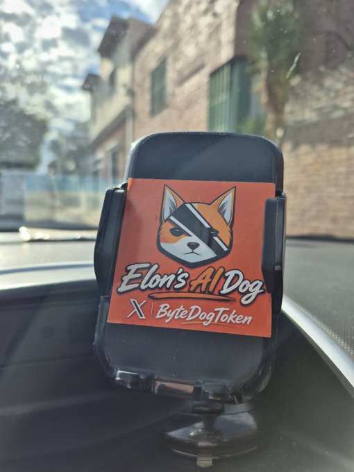 A phone mount on a car dashboard displaying a sticker for 'Elon's AI Dog' promoting the 'ByteDogToken' cryptocurrency with a Shiba Inu logo.