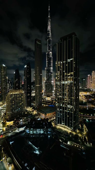 A nighttime aerial view of a city skyline featuring the brightly lit Burj Khalifa towering over surrounding skyscrapers, with illuminated streets visible below.