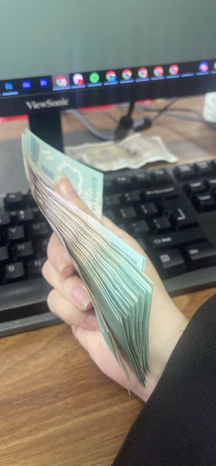 A hand holds a thick stack of Vietnamese đồng banknotes in front of a ViewSonic monitor and keyboard, illustrating the post's message about saving up money from online work and hoping to deposit it into a high-interest bank account after the holiday.