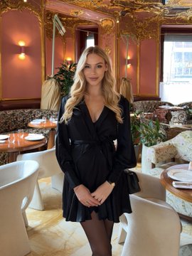 A young blonde woman wearing a black dress with a tied belt stands in an ornately decorated restaurant interior with gold-trimmed pink walls.