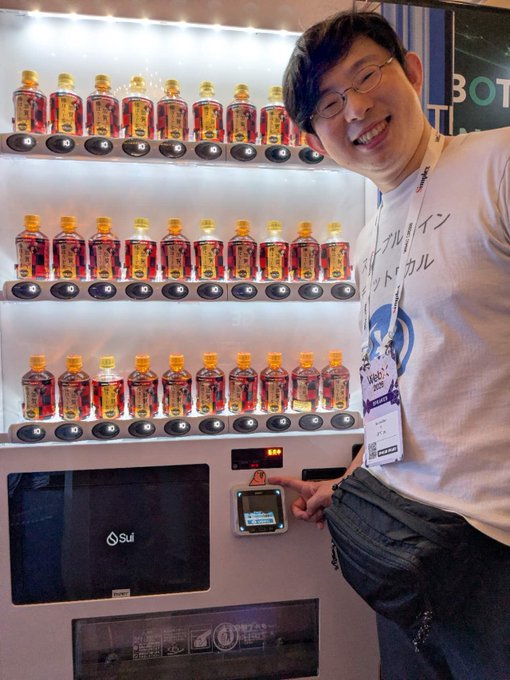Noritaka Okabe, associated with JPYC, points at the interface of a vending machine filled with bottles of Japanese whiskey, likely promoting a WebX conference giveaway or promotion.