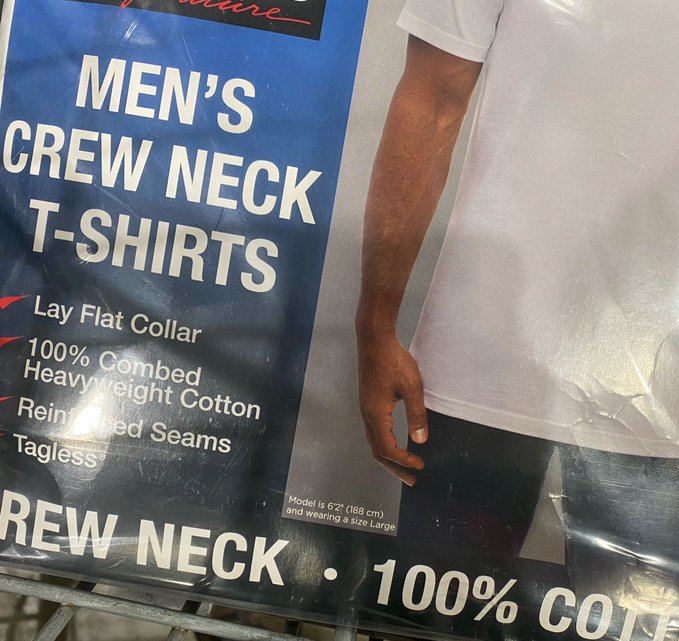 A close-up of a product label reading 'MEN'S CREW NECK T-SHIRTS' with features like 'Lay Flat Collar' and '100% Combed Heavyweight Cotton,' showing a model wearing a white t-shirt and size Large.
