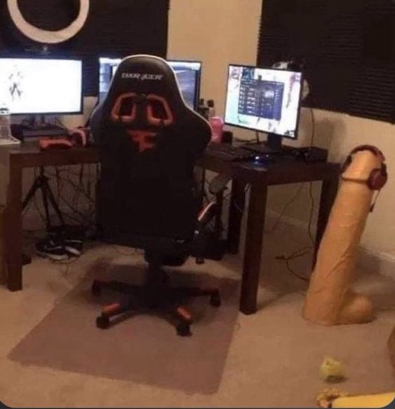 A gaming setup featuring a DXRacer racing-style gaming chair positioned in front of three monitors on a desk, with an unusual tan-colored cylindrical object placed on the floor beside the chair in a room with sound-dampening panels.