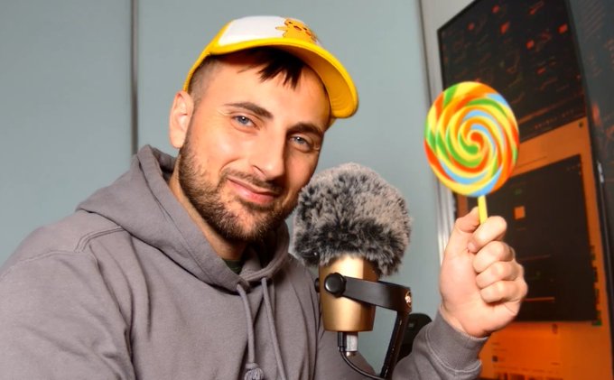A man wearing a yellow cap and gray hoodie smiles while holding up a colorful swirled lollipop, with a professional microphone visible in the foreground, suggesting a podcast or streaming setup. The playful "crypto candy" vibe ties into his post about liking crypto, possibly hinting at a lighthearted take on the market.