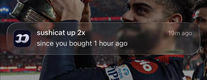 A notification from a crypto trading app showing a user named 'sushicat up 2x' with a message 'since you bought 1 hour ago,' overlaid on an image of a triumphant athlete celebrating with a trophy.