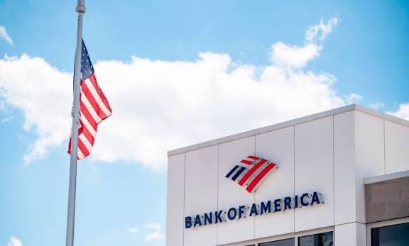 A photograph of a Bank of America building with its red, white, and blue flag logo on the facade, accompanied by an American flag flying at half-mast against a partly cloudy blue sky.