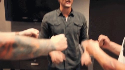 The image shows a group of people standing around each other, with one person holding their fist up in the air while another person has their fist down. The scene appears to be taking place inside a building or room, possibly a living area or a common space where friends gather and socialize. There is also a TV visible in the background, suggesting that this gathering might take place in a home setting with entertainment options available for everyone present.