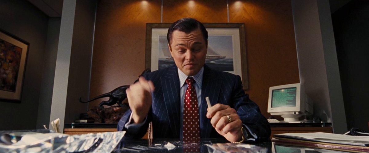 A man wearing a suit, tie, and red scarf sits at his desk with a computer mouse near him as he interacts with it - possibly using an online platform or tool to engage with others or receive information. In front of him on the desk is a pack of cigarettes and a pen. The image also contains text that says "$100 for a comment". It appears this man might be attempting to make a donation in exchange for a comment, possibly related to his recent work activity.