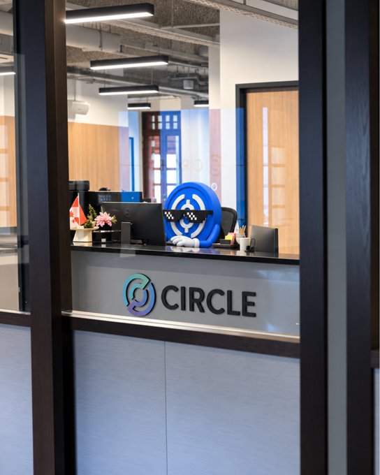 The image shows a Circle company office reception area, featuring a desk with a pixelated "deal with it" sunglasses emoji prop in front of a circular logo. The text "CIRCLE" is prominently displayed on the front of the desk, connecting to the post's announcement of Circle's (@arc) upcoming launch.