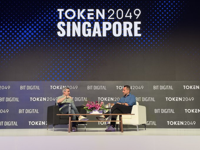 Two men are seated on a stage having a discussion at the 'TOKEN 2049 SINGAPORE' event, with the backdrop displaying 'TOKEN 2049 SINGAPORE' and sponsor logos including 'BIT DIGITAL'.