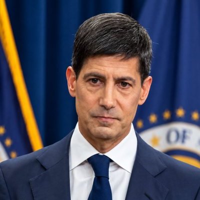 A portrait of a man in a navy suit and blue tie, standing in front of a blue curtain and a partial seal with 'OF' visible.