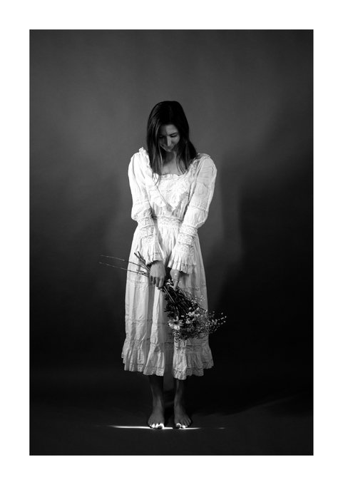 A black-and-white photograph of a barefoot woman in a long white ruffled dress, looking down while holding a bouquet of dried flowers against a dark backdrop.