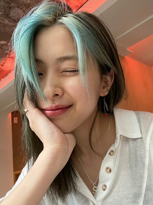 A young woman with mint green and dark hair winks and smiles, resting her chin on her hand while wearing a white button-up shirt.