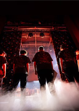 Four men viewed from behind, walking through white stage smoke toward a brightly lit stage with confetti falling, illuminated by dramatic red lighting.