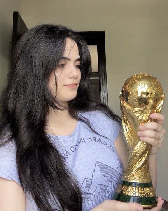 A young woman with long dark hair, wearing a light purple 'ESCAPE IN A DREAM' t-shirt, looks at the FIFA Women's World Cup trophy she is holding.
