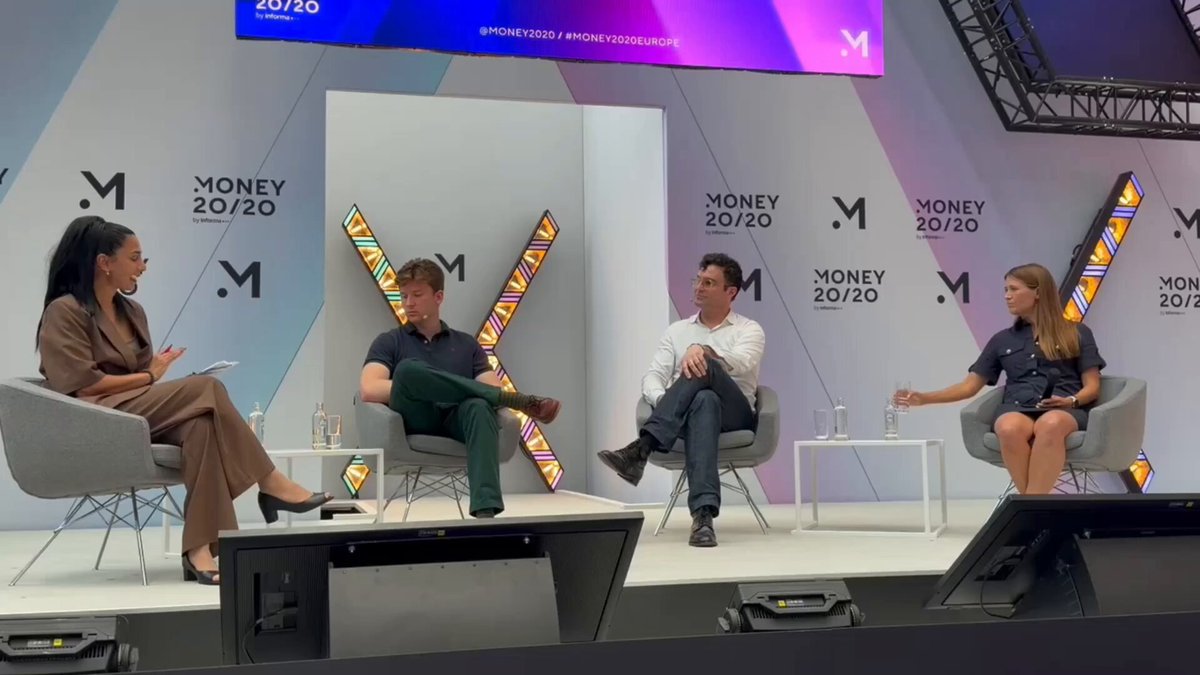 The image shows a group of people sitting around a table at an event, likely discussing privacy concerns related to blockchain technology. The panel consists of three individuals seated on chairs facing each other, with one person standing behind them. They are engaged in conversation about the challenges and benefits of using private balances for financial transactions. The backdrop features a large screen displaying the words "MONEY 2020" along with an image of a white wall adorned with purple and blue stripes.