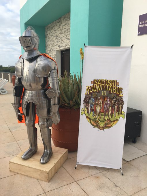 A suit of silver knight armor stands on a wooden pedestal next to a banner for 'Satoshi Round Table III' outside a building.