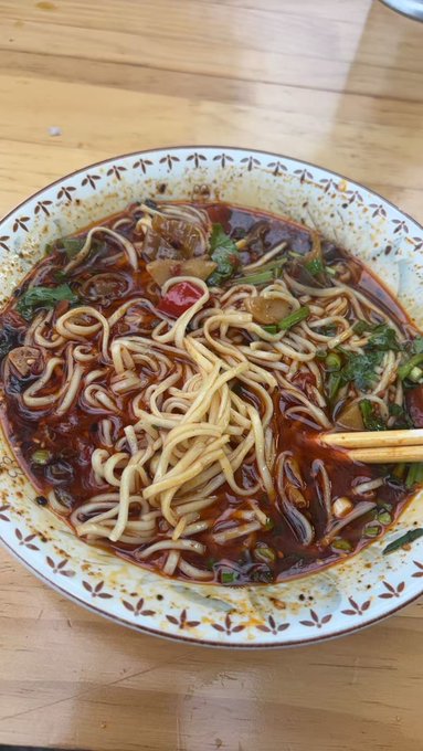 A bowl of spicy red-broth noodles topped with chili oil, green onions, and cilantro sits on a wooden table, with chopsticks resting in the bowl.