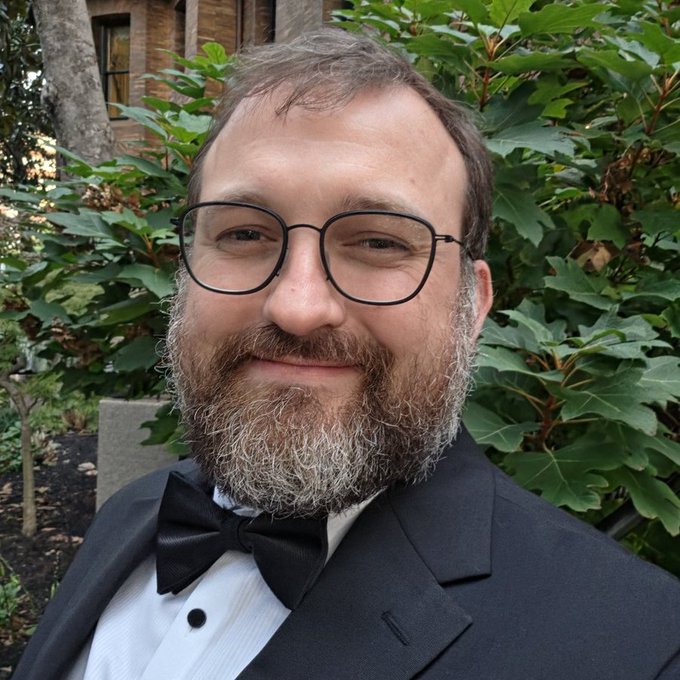 A man with glasses, a beard, and a black tuxedo with bow tie smiles in front of green foliage, appearing ready for the formal "Future of Money, Governance, and the Law" keynote event in DC.