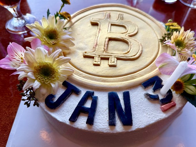 A Bitcoin-themed birthday cake decorated with a large gold Bitcoin logo and "JAN" letters in blue, surrounded by fresh flowers and a lit candle. The cake humorously ties into the birthday wish by featuring Bitcoin imagery, suggesting a celebration paired with hopes for the cryptocurrency's price to soar to $1 million.