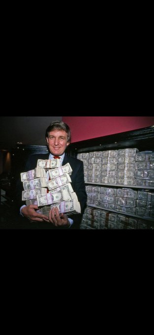 A man in a suit holding a large stack of bundled US currency, standing in front of a wall filled with additional stacks of money.