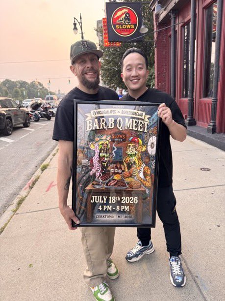 Two men pose on a sidewalk holding a 'Bar BQ Meet' event poster for July 18th, 2026 at Slows Bar BQ in Corktown, Detroit, with a Slows sign visible behind them.