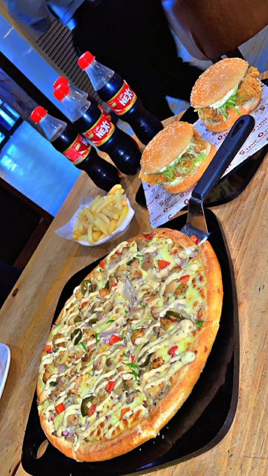 A meal featuring a large oval flatbread/pizza topped with meat, peppers, olives, and sauce, accompanied by two burgers, fries, and three bottles of 'XEX' soda on a wooden table.