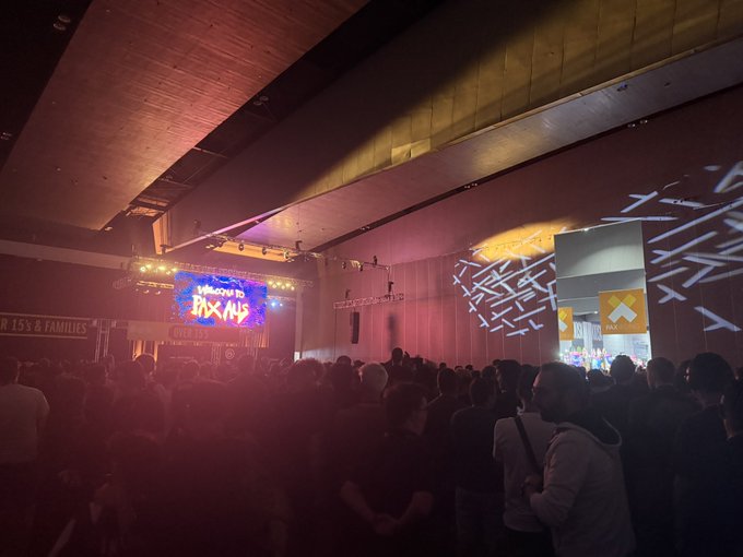 A crowded indoor event space with dim lighting and pink/purple stage lights illuminates a stage displaying 'Welcome to the Box' on a screen, with attendees gathered in the foreground and a textured wall featuring light projections on the right.