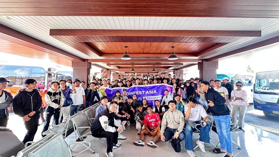 A large group of people poses together under a covered bus terminal, holding a purple banner that reads 'COINFEST ASIA YOGYAKARTA,' with several buses parked nearby.