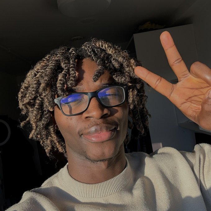 A young man with curly hair and glasses wearing a cream sweater makes a peace sign in a selfie taken indoors with natural sunlight.