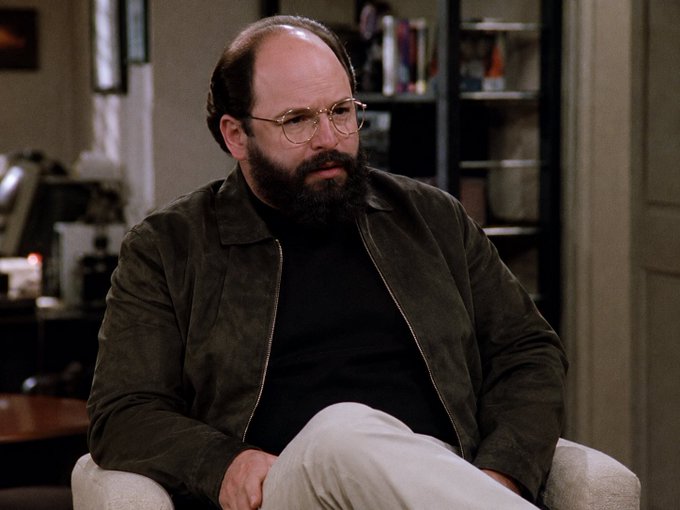 A balding, bearded man wearing glasses and a brown suede jacket over a black turtleneck sits in a chair in an apartment setting.