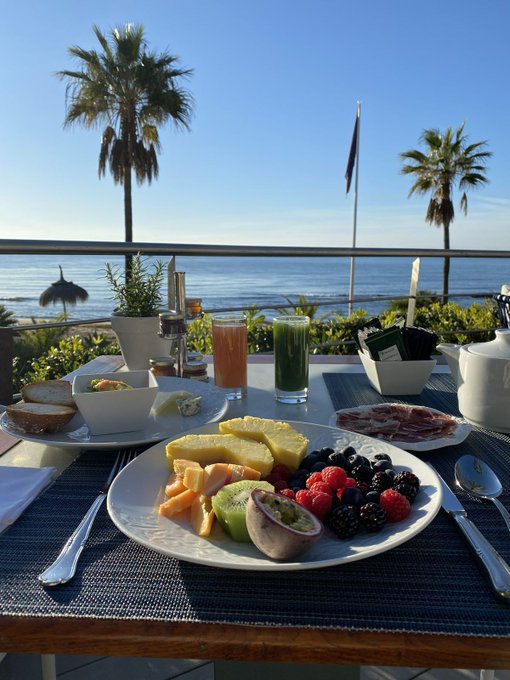 A breakfast spread on a balcony overlooking the ocean, featuring a plate of fresh fruit including pineapple, kiwi, passion fruit, berries, and melon, alongside bread, cheese, cured meats, and glasses of juice and green tea.