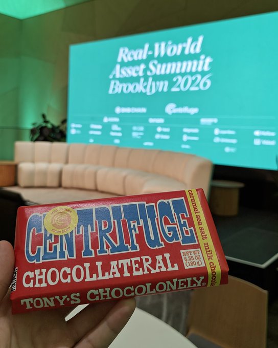 A hand holds a red "TONY'S CHOCOLONELY" candy bar labeled "CENTRIFUGE CHOCOLLATERAL" in front of a screen displaying "Real-World Asset Summit Brooklyn 2026."