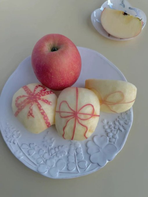 A white decorative plate holds a whole red apple alongside three peeled apples decorated with red lines to resemble wrapped gift boxes, with another apple slice on a smaller plate behind it.
