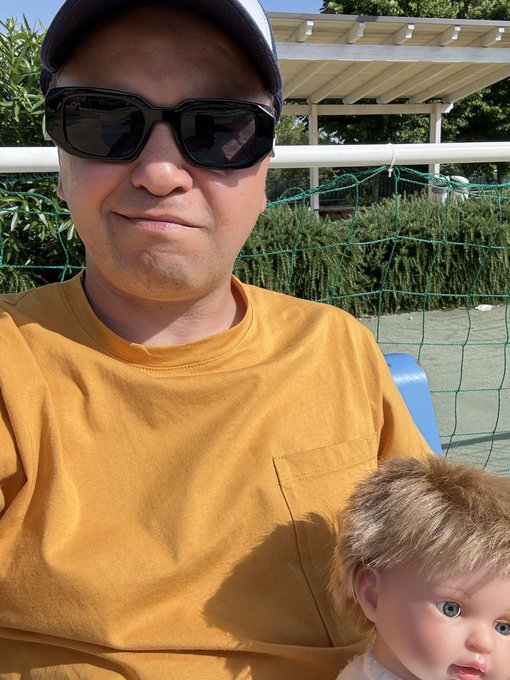 A selfie of a man wearing sunglasses and a yellow shirt holding a doll, taken outdoors near a tennis court.