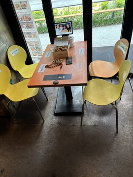 The image shows a dining area with four chairs placed around an orange table. There are two yellow chairs, one blue chair, and one green chair. On top of the table, there is a laptop computer along with some snacks such as cookies and crackers. The scene appears to be set in a restaurant or cafe where people can enjoy their meals while watching TV shows on a large screen television mounted on the wall.