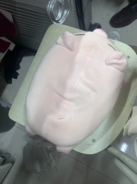 A pink, round plush toy resembling a pig or hamster is shown from above, resting on a green surface.