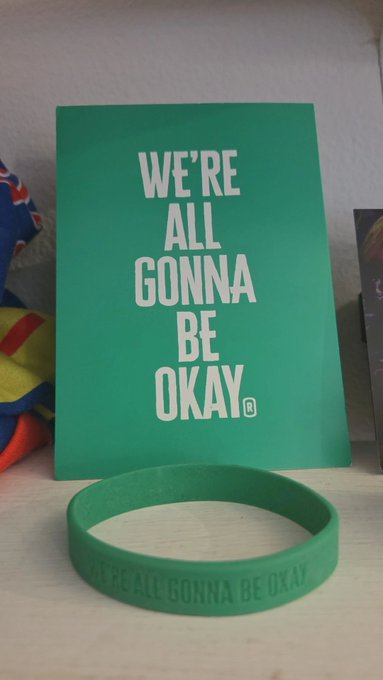 A green greeting card displaying the text 'WE'RE ALL GONNA BE OKAY®' placed above a matching green silicone wristband with the same message engraved on it.