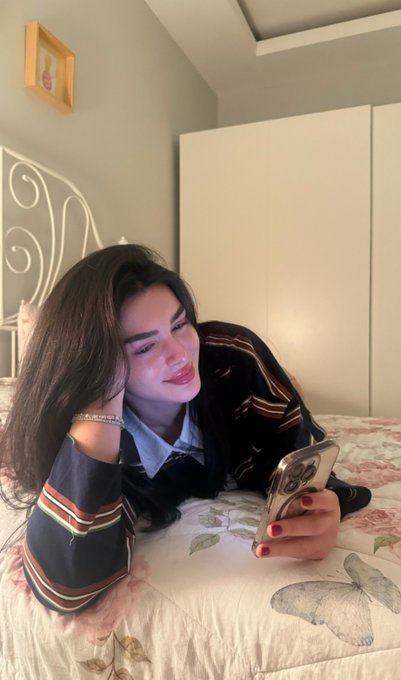 A young woman with long dark hair lies on a floral bedspread, smiling down at her smartphone while wearing a striped sweater and bracelet.