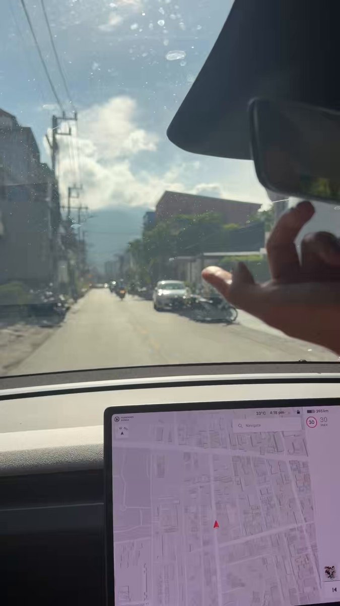 This image captures a moment where the driver of a car is navigating through an area using a GPS device placed on their dashboard, which is positioned just below the windshield mirror. The scene outside the window features multiple cars driving along the road with buildings visible in the background. The caption provided about Thailand and university leadership adds context to this picture by talking about university support for the director's company, and the additional hardware devices purchased last month. It seems that the driver is taking a break from their journey and observing the surroundings or possibly planning their next move based on the GPS directions given by the device.