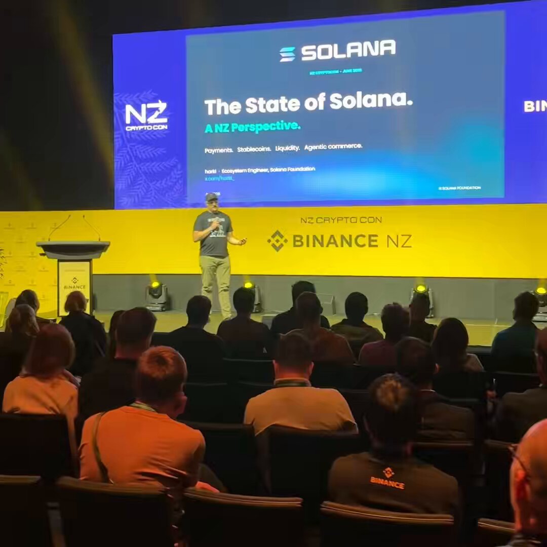 The image shows a man standing at a podium, giving a presentation or speech to a group of people seated in chairs behind him. The audience appears engaged and attentive as they listen to his words. The background features a large screen displaying information related to Solana, which is likely part of the presentation.