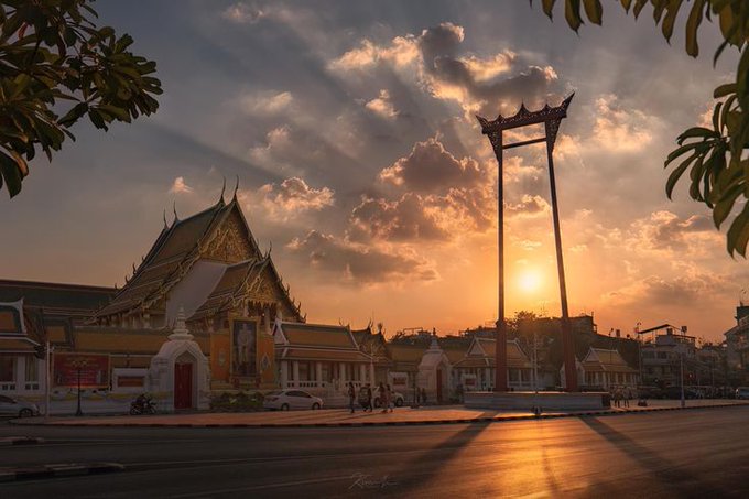 Scenic sunset photograph of a Thai temple and the Giant Swing (Sao Ching Cha) in Bangkok, with golden clouds and silhouetted foliage.